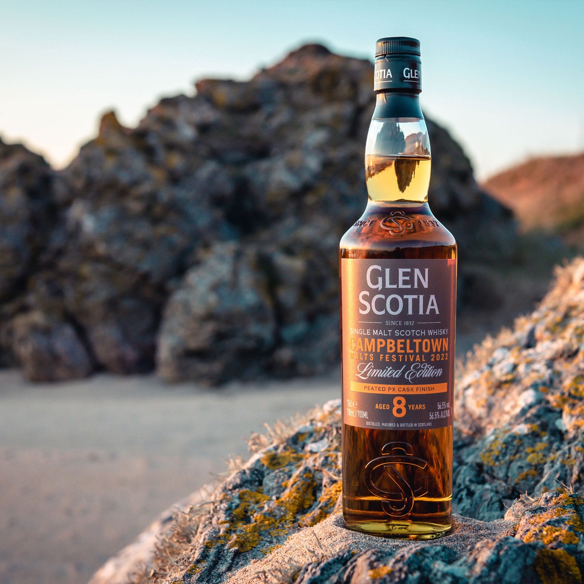 A bottle of Glen Scotia Campbeltown Malt Scotch Whisky sits on a rocky surface outdoors. The label indicates it is an 8-year-old Limited Edition for the Campbeltown Malts Festival 2021. A serene landscape with blurred natural elements is visible in the background.