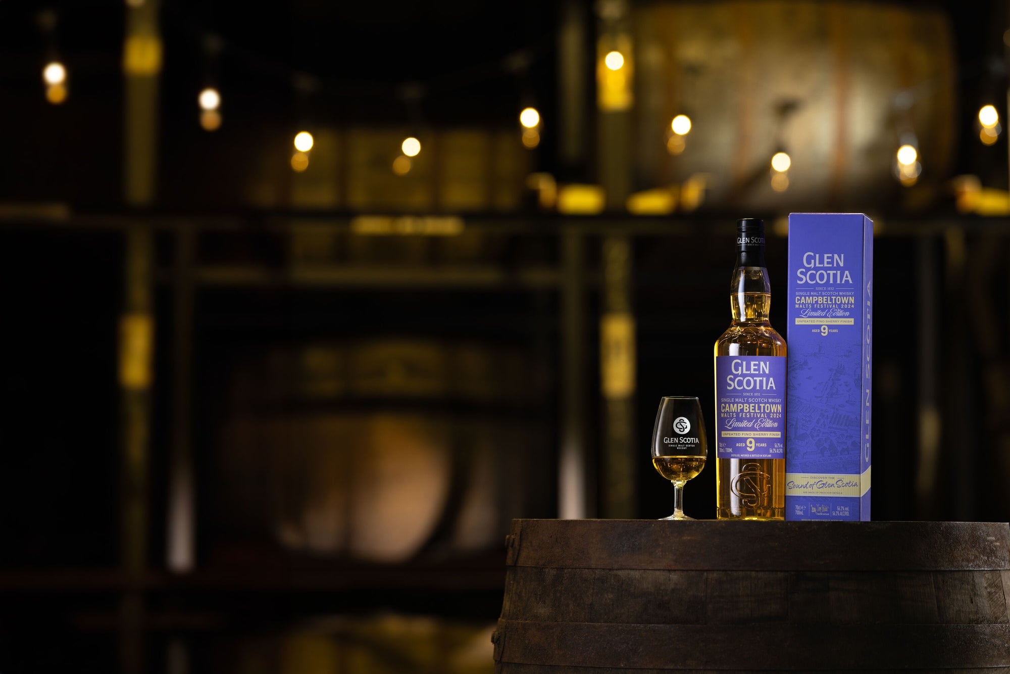 A bottle of Glen Scotia Campbeltown 9-year-old scotch whisky and its box are placed on a wooden barrel. A branded Glen Scotia whisky glass is also on the barrel. The background features dim lighting with string lights and large wooden barrels.