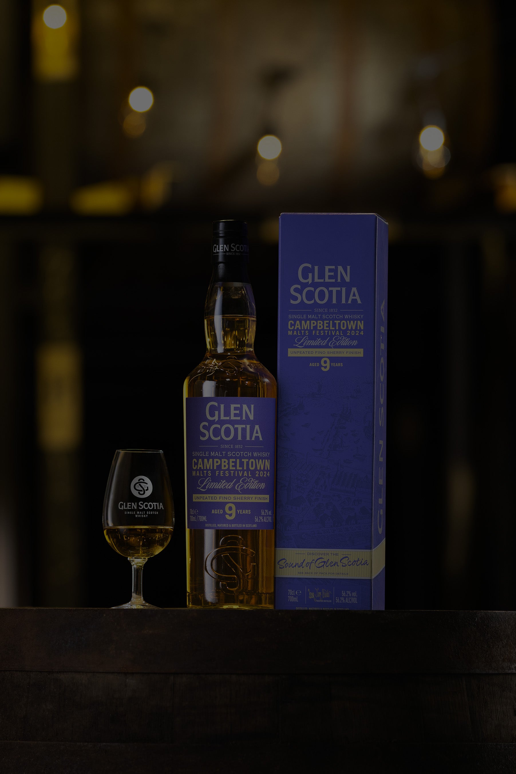 A dimly lit image showcasing a bottle of Glen Scotia Campbeltown 9-year-old single malt scotch whisky, next to its blue and gold box. A Glen Scotia branded whisky glass is placed nearby, all set against a dark, blurred background.