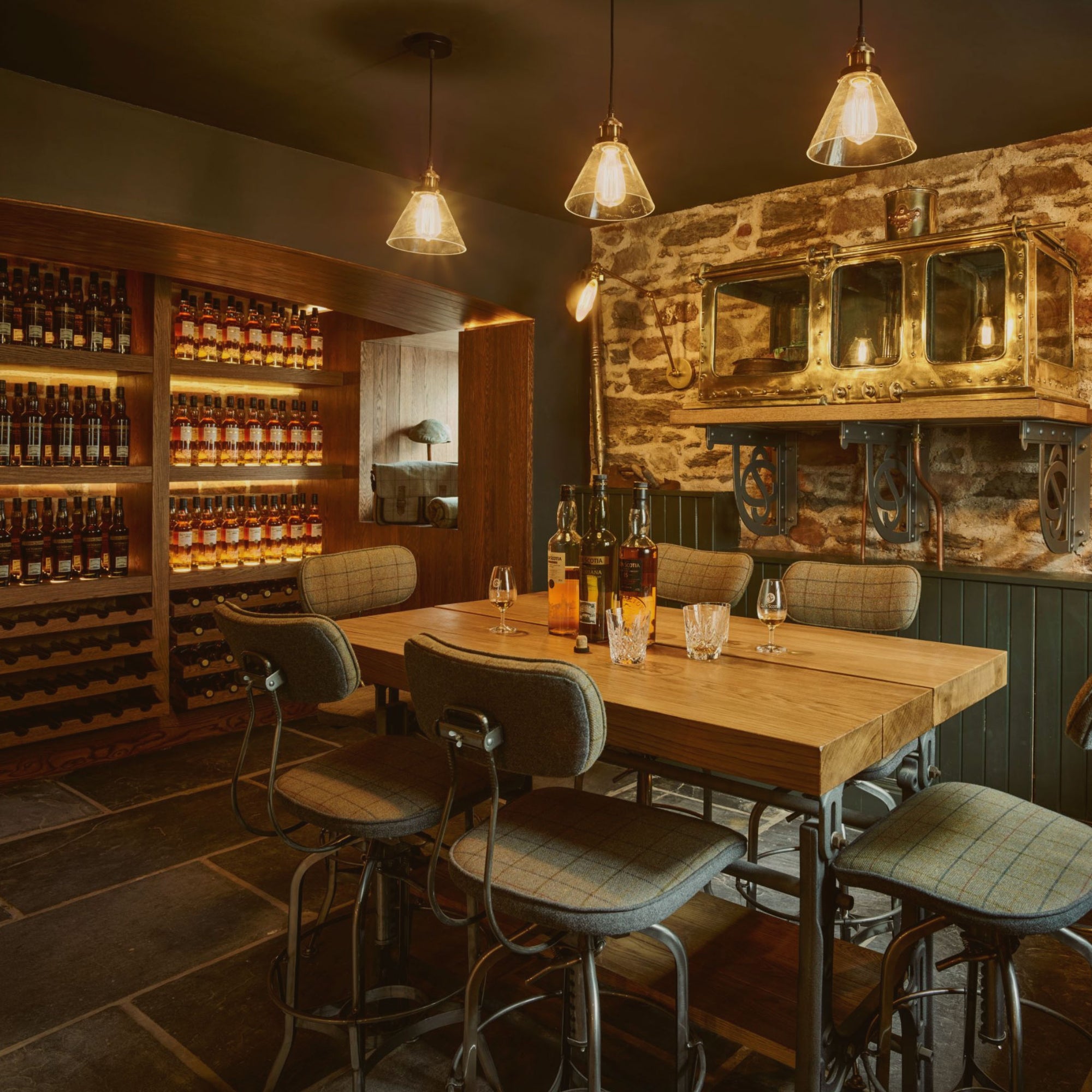 A cozy, dimly lit wine cellar features a wooden table with six cushioned chairs. Shelves on the left are stocked with wine bottles, and the stone wall on the right display antique mirrors and light fixtures above a small counter. Three pendant lights hang overhead.