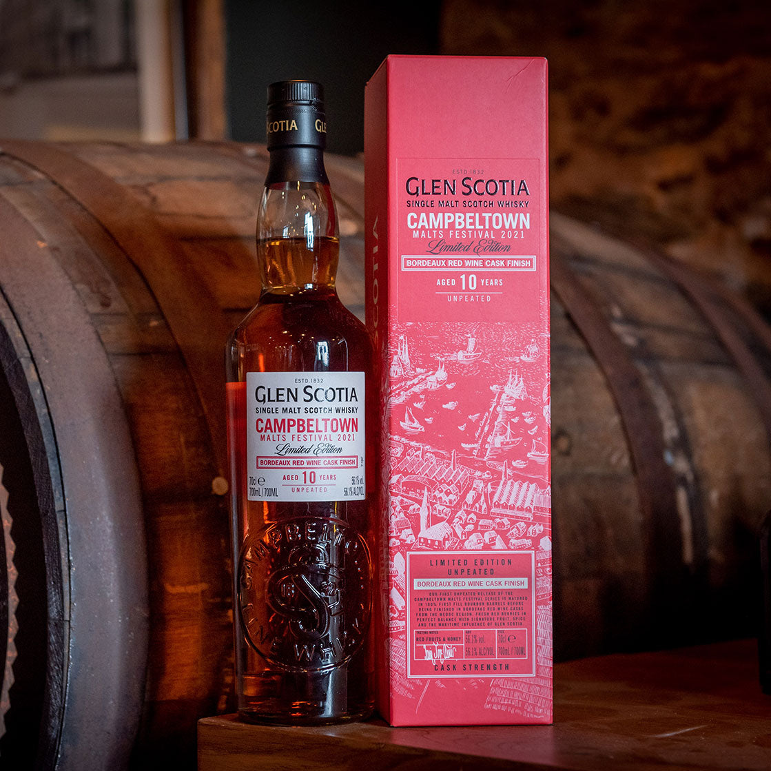 A bottle of Glen Scotia single malt scotch whisky stands next to its red box packaging, labeled "Campbeltown Malts Festival 2021," placed on a rustic wooden barrel background. The whisky is aged 10 years and the packaging includes intricate artwork.