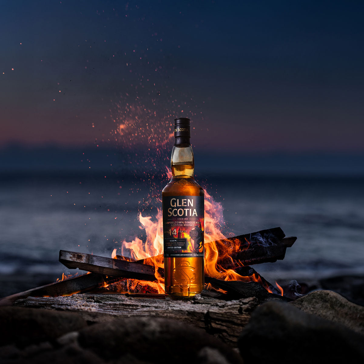 An Icons of Campbeltown Release No.2 bottle by Glen Scotia is positioned in front of a crackling fire, with a dark, blurry ocean backdrop at twilight. Embers from the fire float around the bottle, creating a visually striking contrast between the warm flames and the cool, dark surroundings.