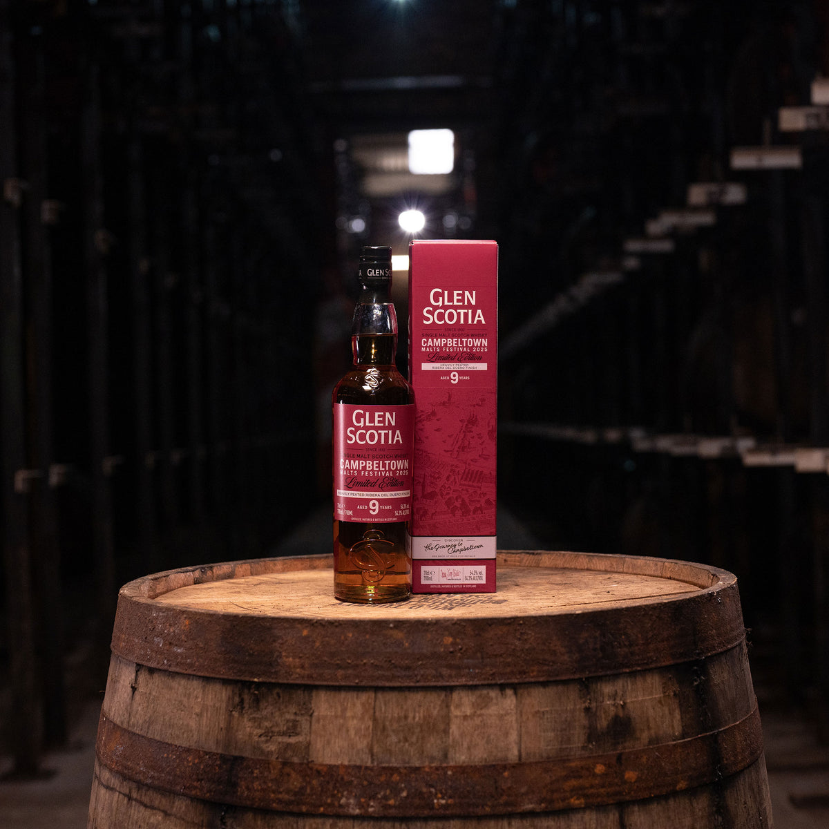 A bottle of Glen Scotia Campbeltown Malts Festival Edition 2025 rests on a wooden barrel next to its box. Finished in Ribera del Duero wine casks, it&#39;s set in a dimly lit cellar with rows of wooden casks, highlighting its unique character.